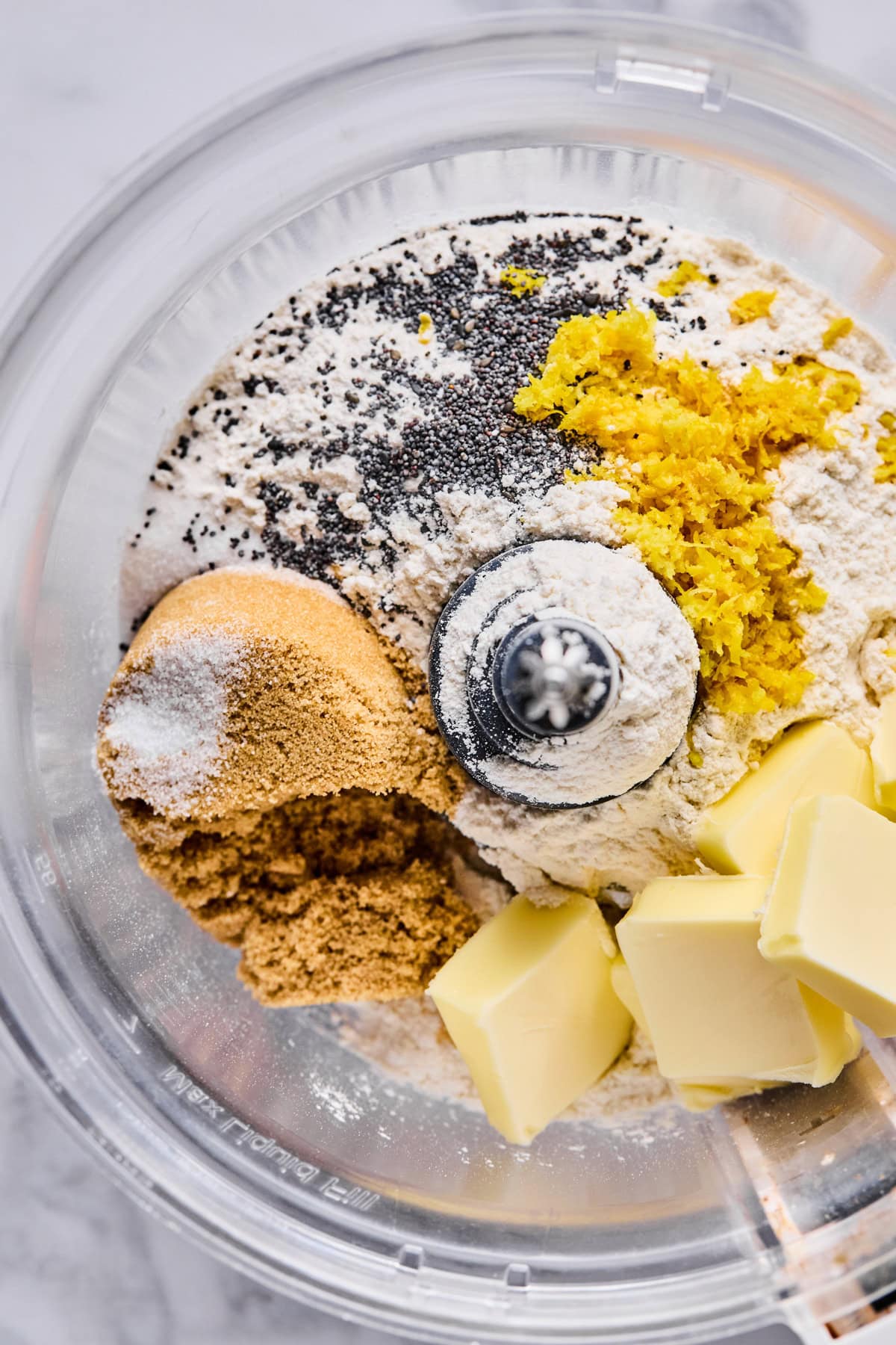 A food processor bowl containing flour, brown sugar, poppy seeds, lemon zest, salt, and cubes of butter, ready to be mixed.