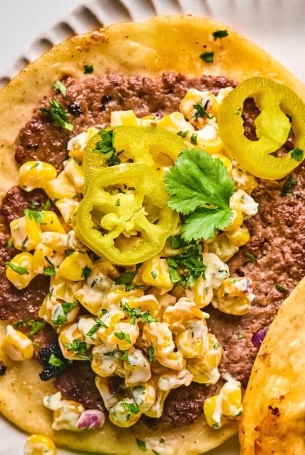 A close-up of a taco with ground beef, creamy corn salad, pickled jalapeño slices, and a sprig of cilantro on a white plate.