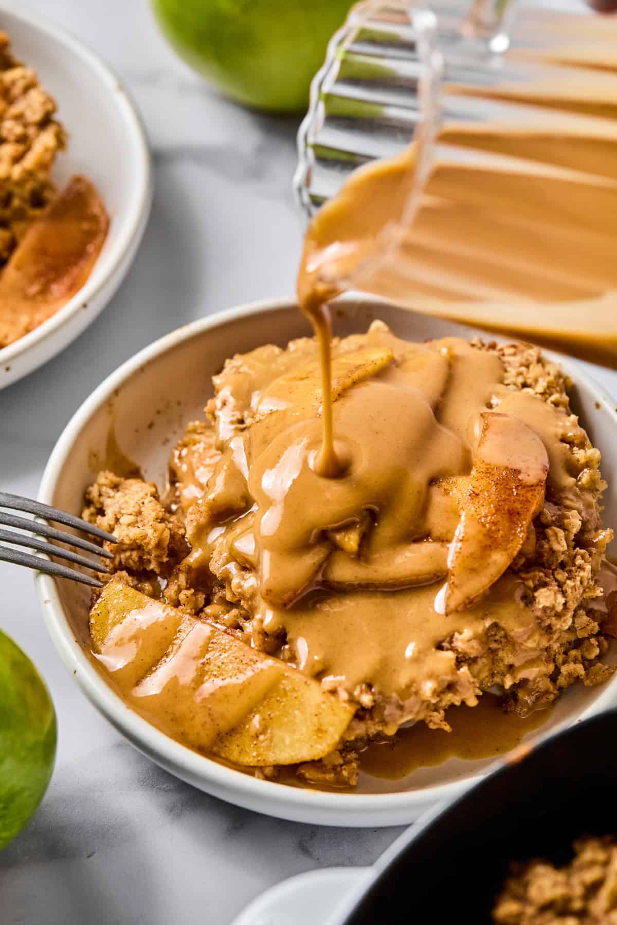 A bowl of apple crisp is topped with caramel sauce being poured from a glass container, with apples visible and a fork resting on the side.