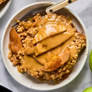 A bowl of apple crisp topped with sliced apples and a generous drizzle of nut butter, with a fork resting in the bowl.