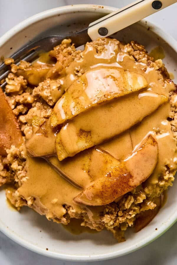 A bowl of apple crisp topped with sliced apples and a generous drizzle of nut butter, with a fork resting in the bowl.