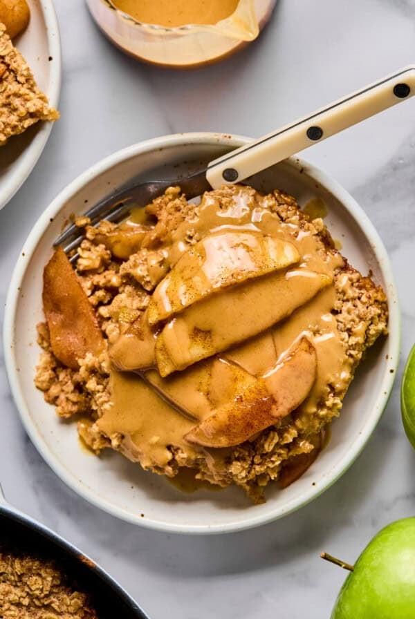 A slice of apple crumble topped with sliced apples and a drizzle of caramel sauce on a white plate with a fork.