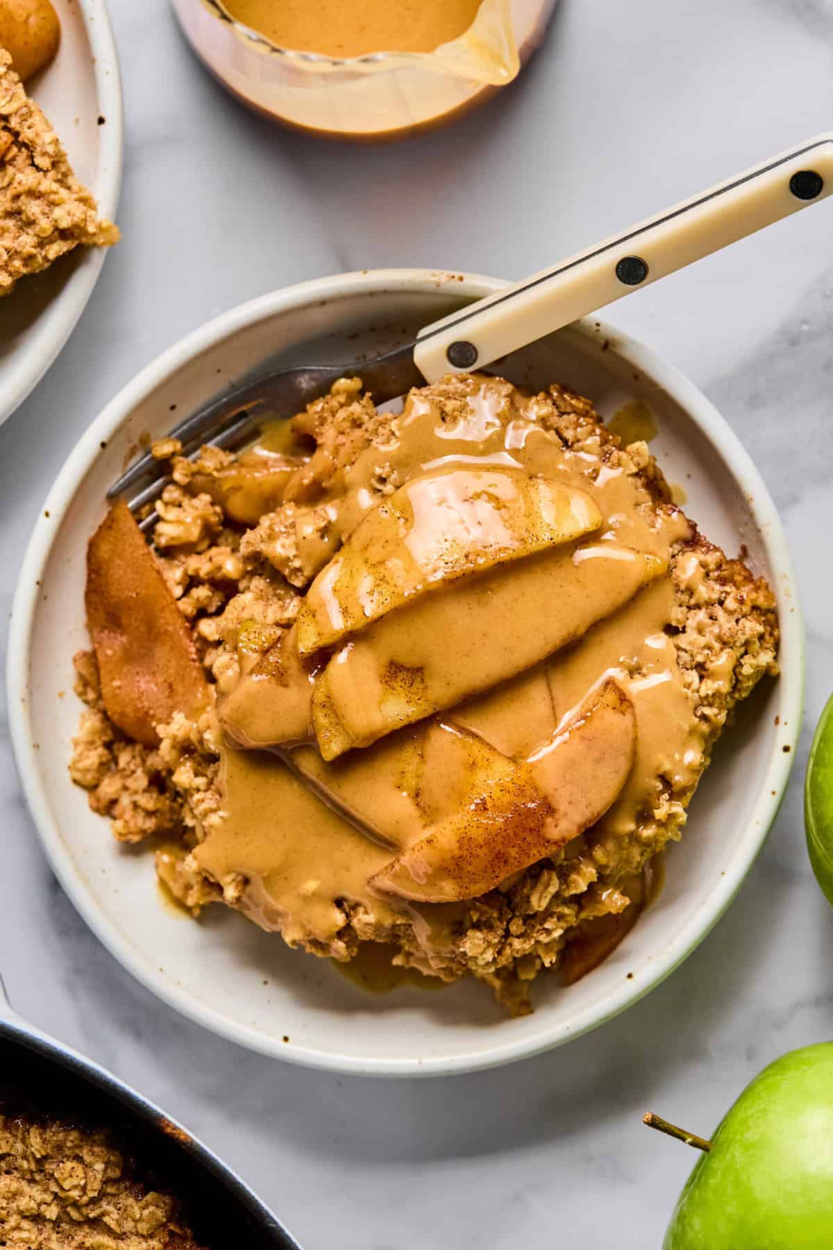 A slice of apple crumble topped with sliced apples and a drizzle of caramel sauce on a white plate with a fork.