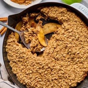 A cast iron skillet filled with baked apple crisp topped with oat crumble, with a serving scooped out onto a plate. Green apples and cinnamon sticks are nearby.