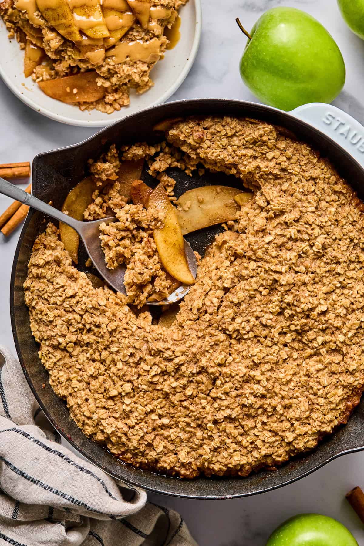 A cast iron skillet filled with baked apple crisp topped with oat crumble, with a serving scooped out onto a plate. Green apples and cinnamon sticks are nearby.