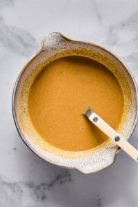 A ceramic bowl filled with a smooth, light brown liquid mixture and a white-handled spoon resting inside, placed on a marble surface.