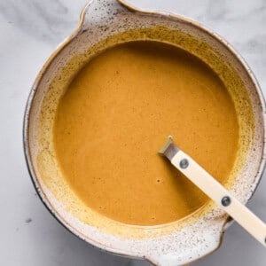 A ceramic bowl filled with a smooth, light brown liquid mixture and a white-handled spoon resting inside, placed on a marble surface.