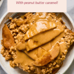 A bowl of apple pie baked oatmeal topped with peanut butter caramel sauce, with a fork on the side and an apple partially visible nearby.