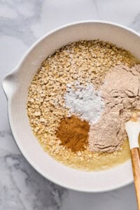 A mixing bowl containing oats, flour, baking powder, cinnamon, and a liquid ingredient with a spatula resting on the side.