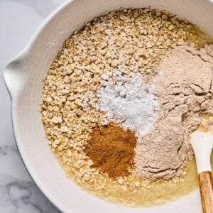 A mixing bowl containing oats, flour, baking powder, cinnamon, and a liquid ingredient with a spatula resting on the side.