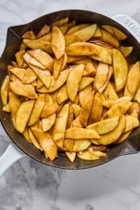 Sliced apples coated with cinnamon and sugar cooking in a cast iron skillet on a marble countertop.
