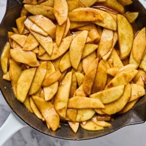Sliced apples coated with cinnamon and sugar cooking in a cast iron skillet on a marble countertop.