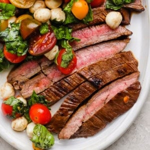 Sliced medium-rare steak served with a salad of cherry tomatoes, mozzarella balls, and fresh basil on a white plate.