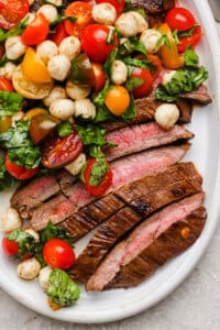 Sliced medium-rare steak served with a salad of cherry tomatoes, mozzarella balls, and fresh basil on a white plate.