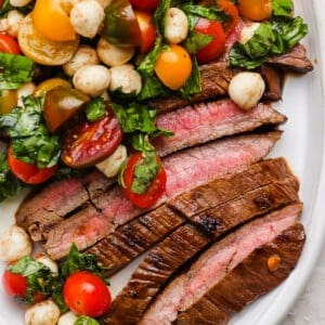Sliced medium-rare steak served with a salad of cherry tomatoes, mozzarella balls, and fresh basil on a white plate.