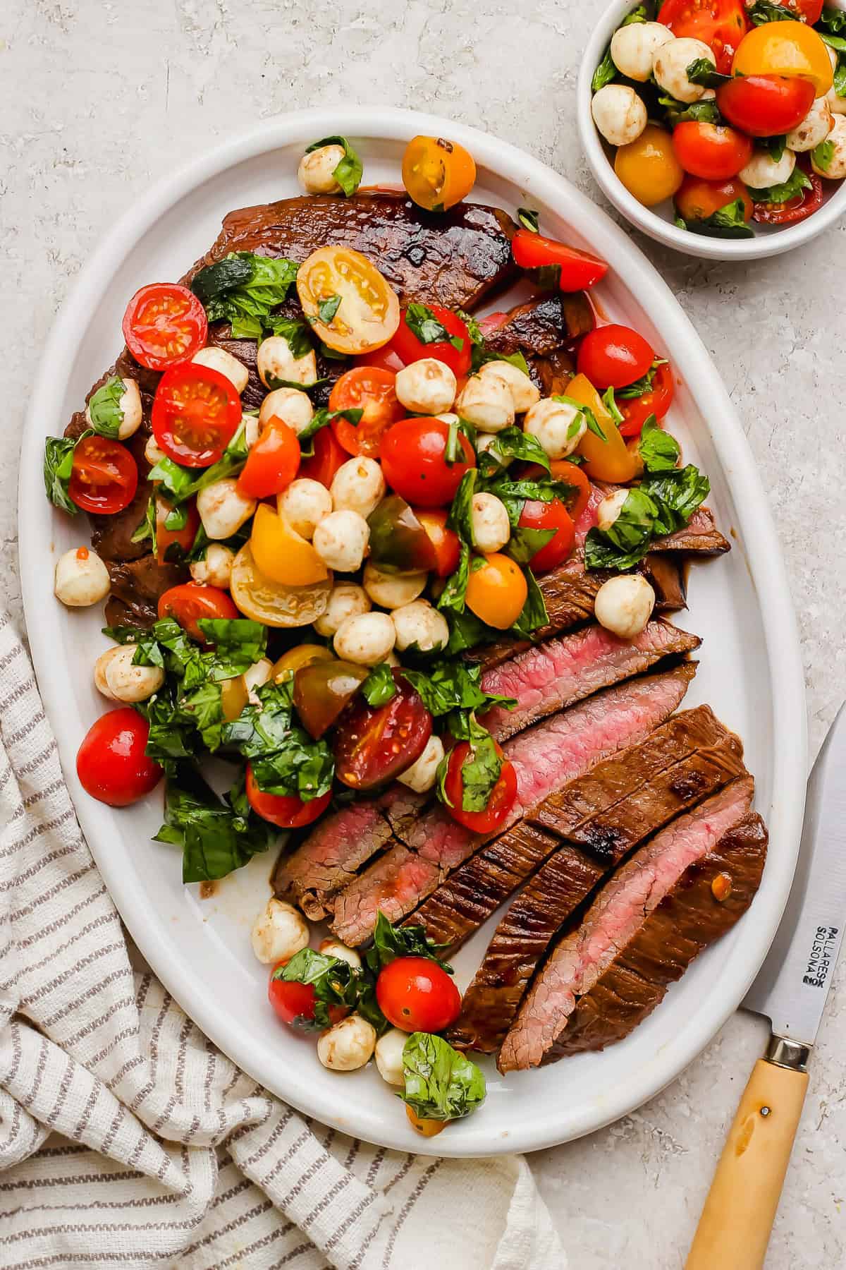 Sliced medium-rare steak on a white oval platter, topped with a tomato, mozzarella, and basil salad; a matching small bowl is beside it. A striped towel and knife are nearby.