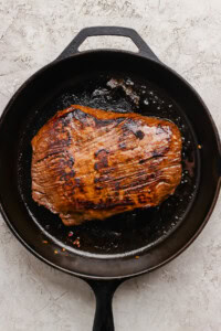 A seared piece of meat is cooking in a black cast iron skillet on a light textured surface.
