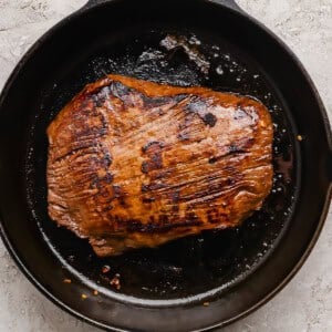 A seared piece of meat is cooking in a black cast iron skillet on a light textured surface.