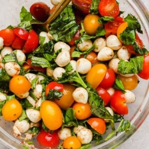A clear glass bowl filled with cherry tomatoes, mozzarella balls, fresh basil leaves, and a spoon, on a light surface.