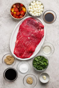 Raw flank steak on a white plate surrounded by bowls of grape tomatoes, mozzarella balls, brown sugar, spinach, balsamic vinegar, Worcestershire sauce, salt, olive oil, mustard, and garlic.