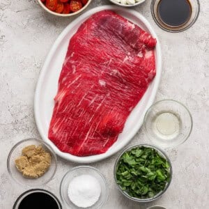 Raw flank steak on a white plate surrounded by bowls of grape tomatoes, mozzarella balls, brown sugar, spinach, balsamic vinegar, Worcestershire sauce, salt, olive oil, mustard, and garlic.