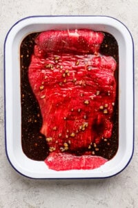 Raw flank steak marinating in a white dish with dark sauce and minced garlic on a light textured surface.