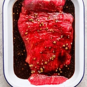 Raw flank steak marinating in a white dish with dark sauce and minced garlic on a light textured surface.
