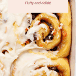 Close-up of carrot cake cinnamon rolls in a baking dish, topped with white icing and visible raisins, with a text overlay describing the dish.