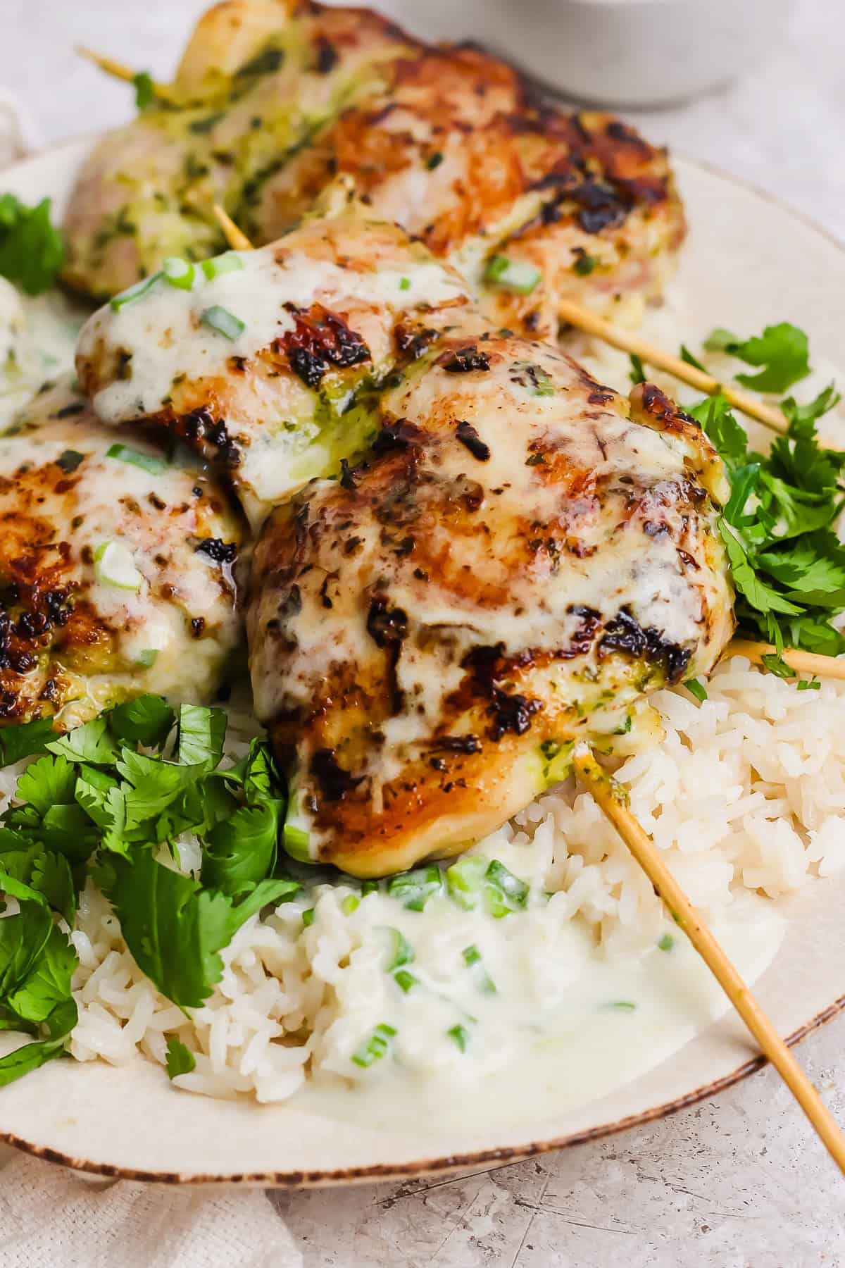 Three grilled chicken skewers topped with a creamy white sauce are served over white rice and garnished with fresh cilantro on a white plate.