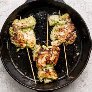 Three grilled, stuffed chicken thighs secured with wooden skewers are cooking in a black cast iron skillet on a light textured surface.