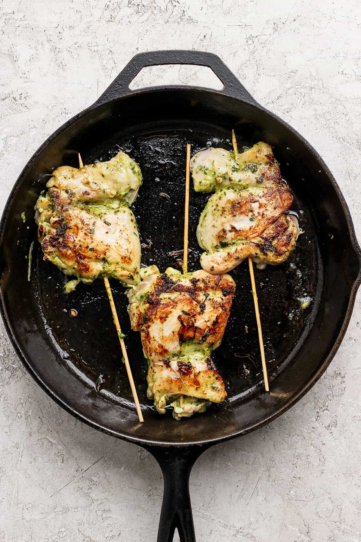 Three grilled, stuffed chicken thighs secured with wooden skewers are cooking in a black cast iron skillet on a light textured surface.