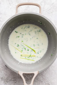 A saucepan containing a creamy white sauce with chopped green herbs and several stalks of lemongrass on a light textured surface.