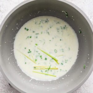 A saucepan containing a creamy white sauce with chopped green herbs and several stalks of lemongrass on a light textured surface.