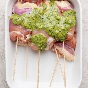 Raw chicken breasts skewered on wooden sticks in a white baking dish, topped with a green herb sauce.