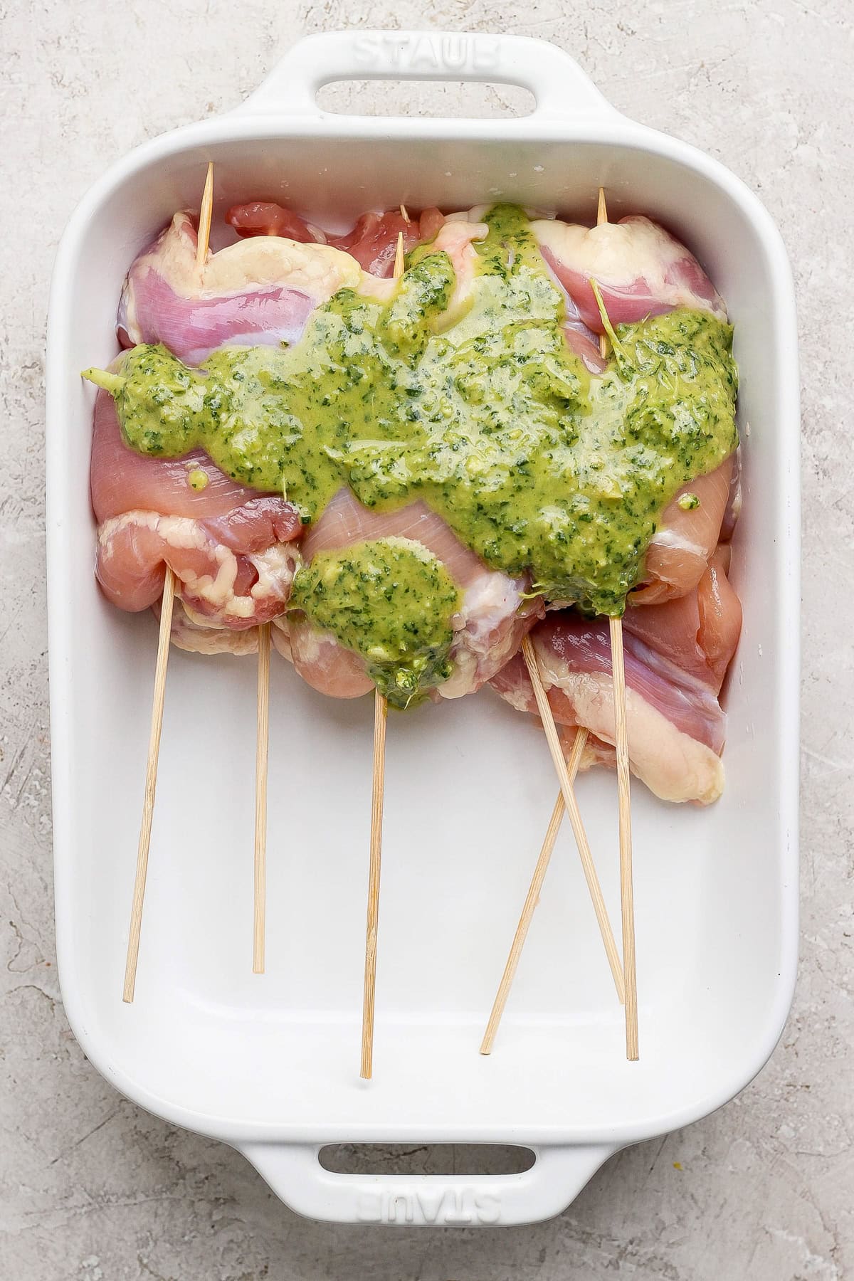 Raw chicken breasts skewered on wooden sticks in a white baking dish, topped with a green herb sauce.