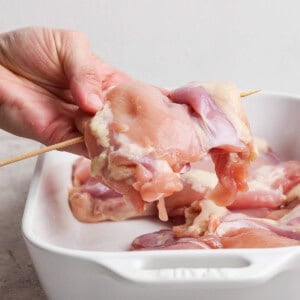 A hand holding raw chicken piece on a wooden skewer over a white ceramic baking dish with more raw chicken pieces inside.