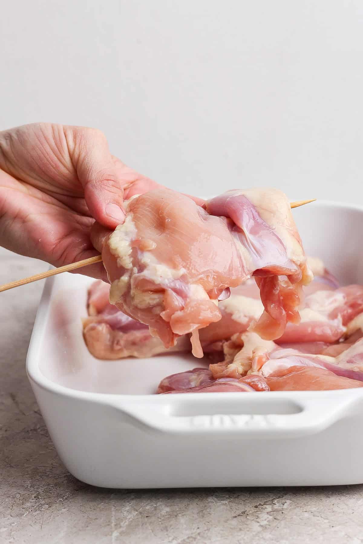A hand holding raw chicken piece on a wooden skewer over a white ceramic baking dish with more raw chicken pieces inside.