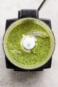 Overhead view of a food processor containing a green sauce or mixture with visible herbs on a light countertop.