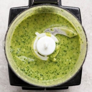 Overhead view of a food processor containing a green sauce or mixture with visible herbs on a light countertop.