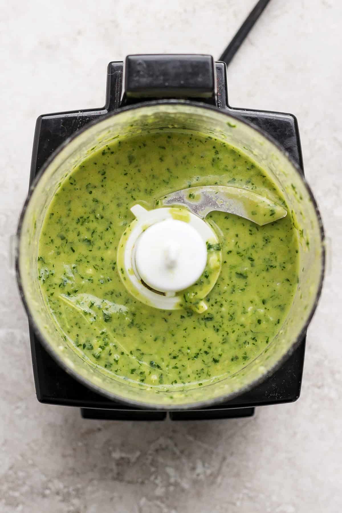 Overhead view of a food processor containing a green sauce or mixture with visible herbs on a light countertop.