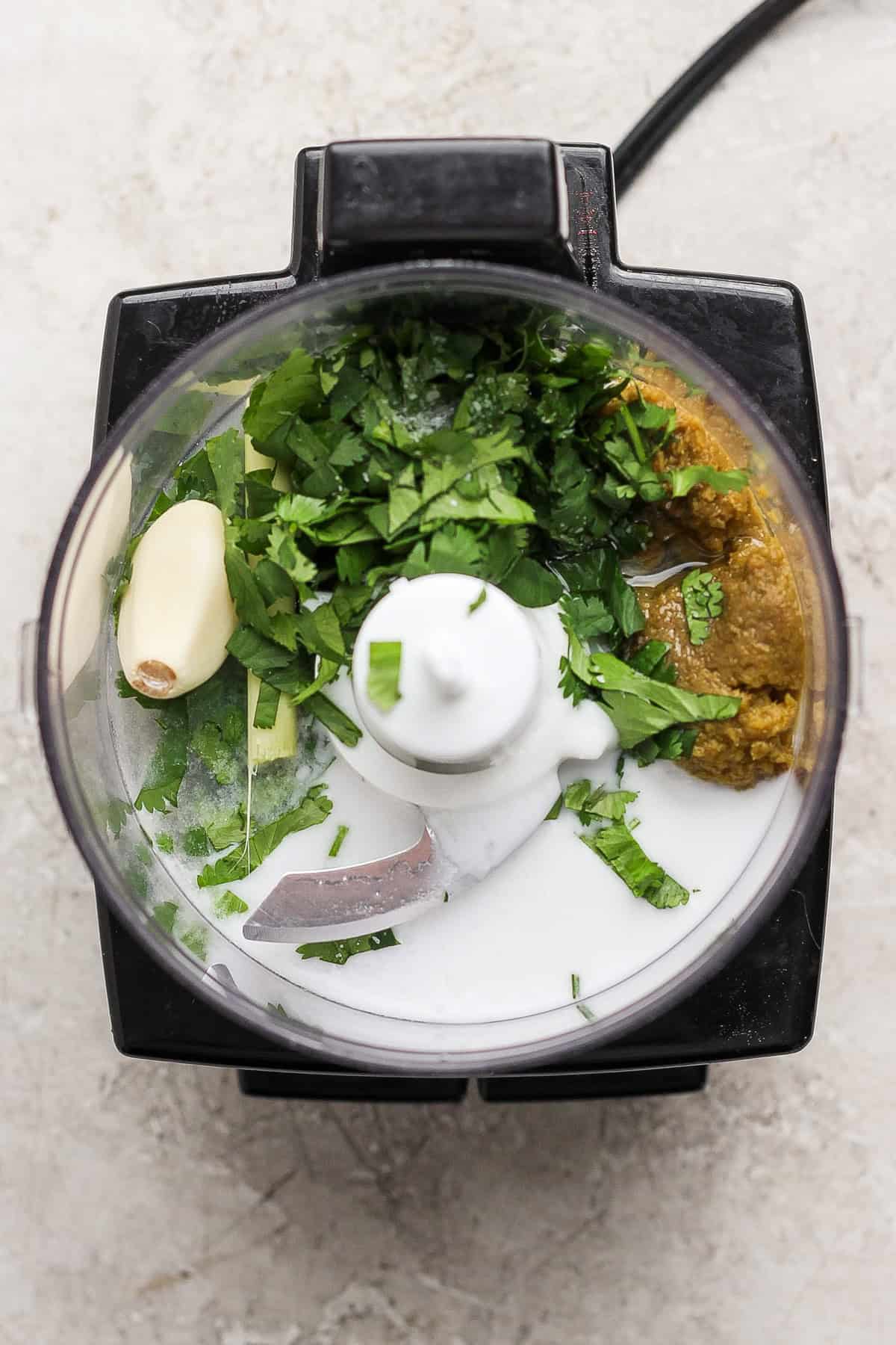 Overhead view of a food processor containing coconut milk, fresh cilantro, garlic, ginger, and a greenish paste, ready to be blended.