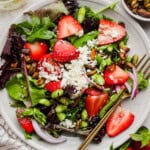 A plate of mixed green salad topped with sliced strawberries, feta cheese, red onion, pistachios, asparagus, and a gold fork on the side.
