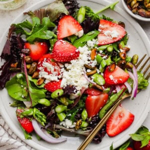 A plate of mixed green salad topped with sliced strawberries, feta cheese, red onion, pistachios, asparagus, and a gold fork on the side.