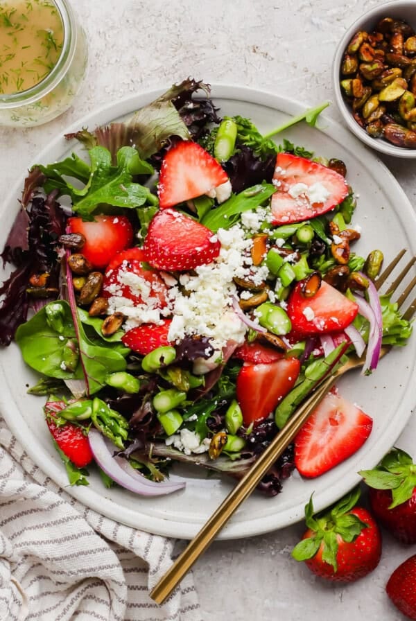 A plate of mixed greens salad with sliced strawberries, crumbled cheese, pistachios, red onion, and a fork, next to a bowl of nuts and fresh strawberries.