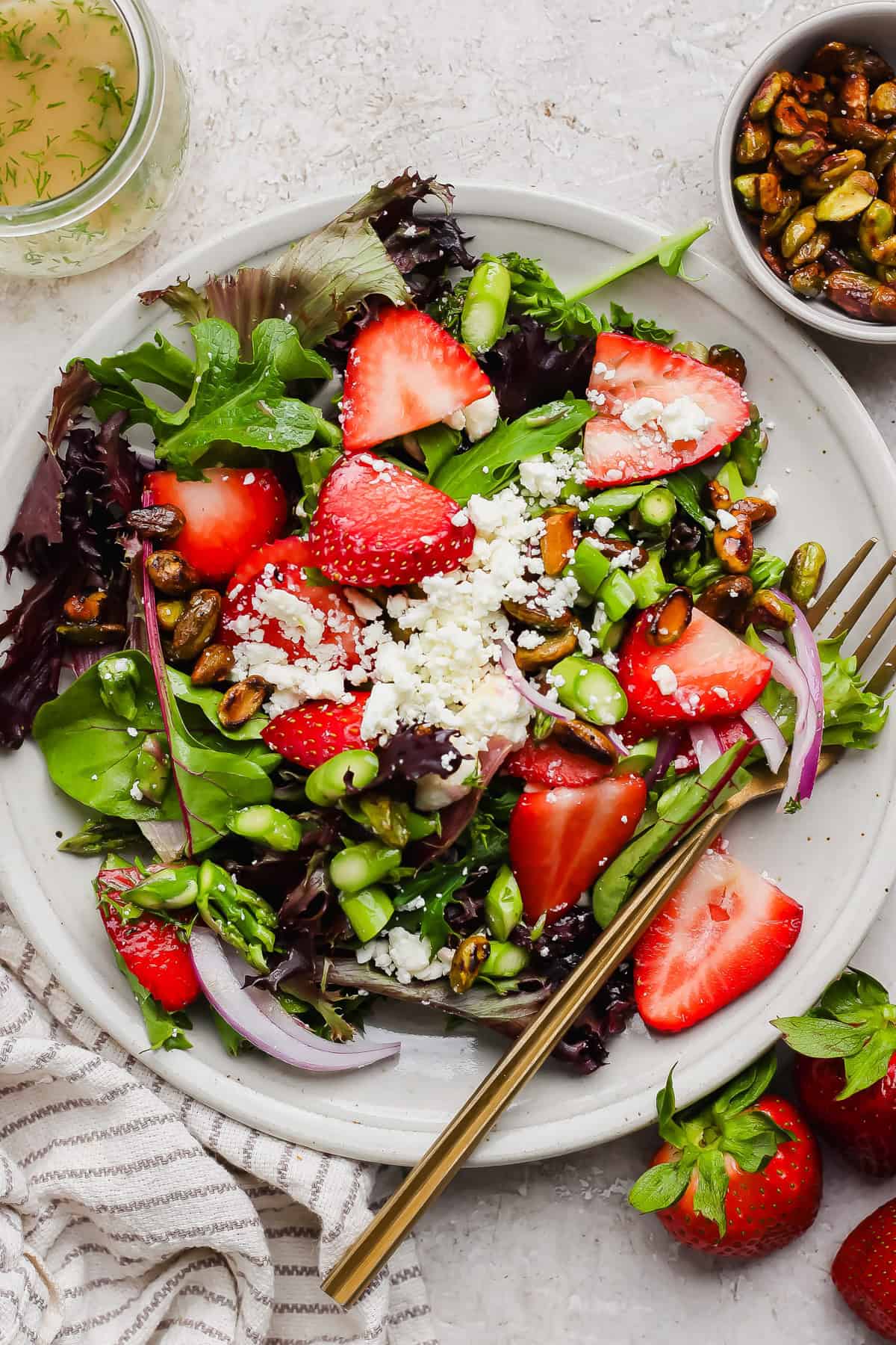 A plate of mixed greens salad with sliced strawberries, crumbled cheese, pistachios, red onion, and a fork, next to a bowl of nuts and fresh strawberries.