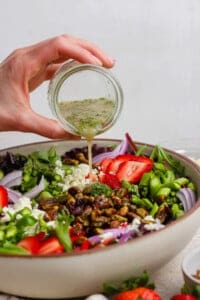 A hand pours vinaigrette from a small jar onto a salad with strawberries, feta cheese, red onions, pistachios, and greens in a large bowl.