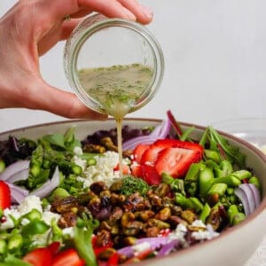A hand pours vinaigrette from a small jar onto a salad with strawberries, feta cheese, red onions, pistachios, and greens in a large bowl.