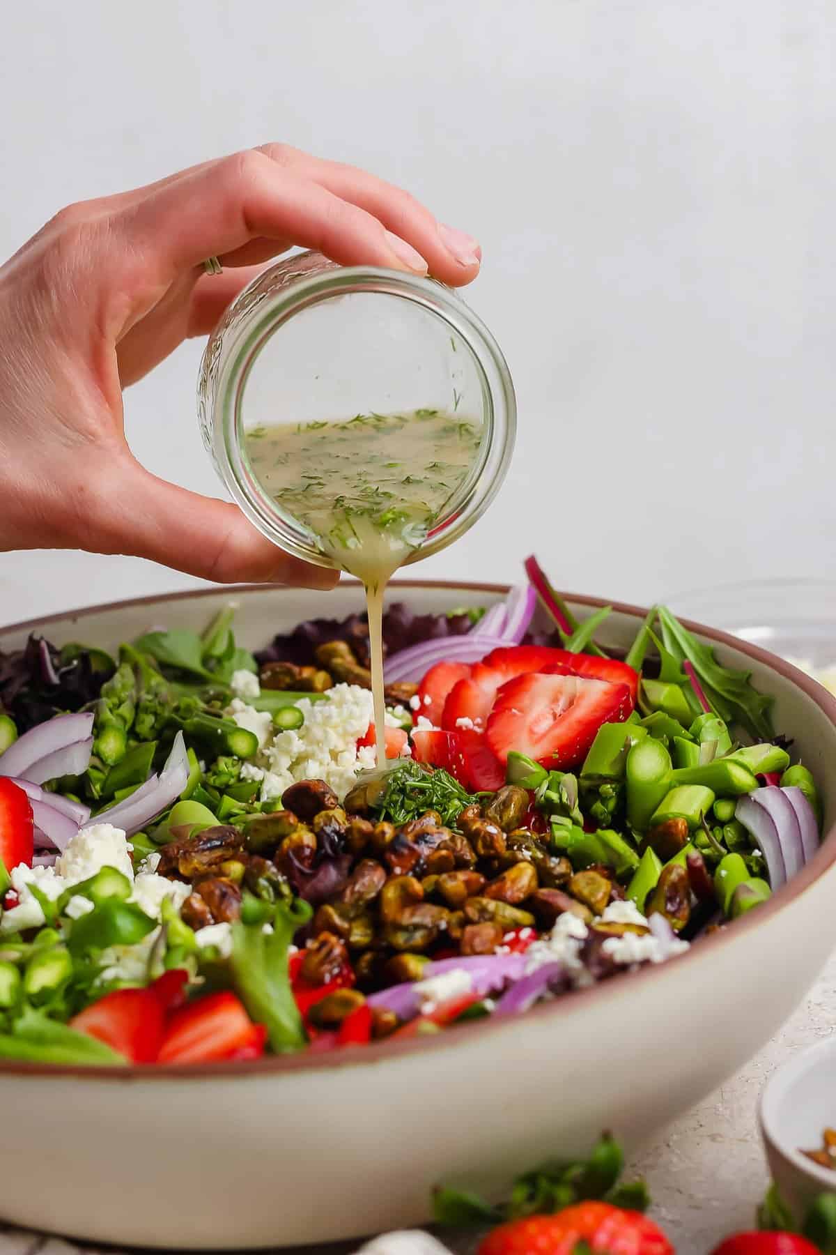 A hand pours vinaigrette from a small jar onto a salad with strawberries, feta cheese, red onions, pistachios, and greens in a large bowl.