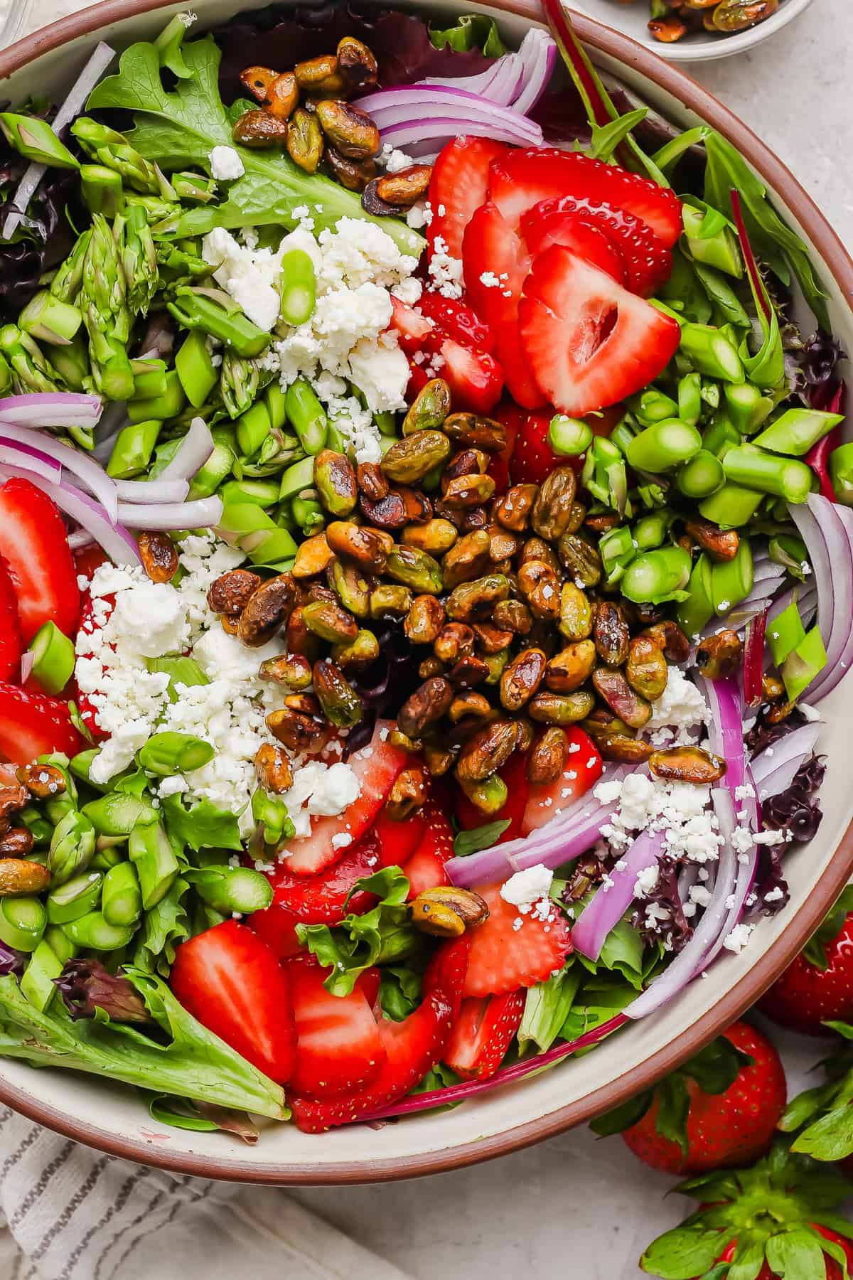 A salad with mixed greens, sliced strawberries, red onions, asparagus, edamame, crumbled cheese, and roasted pistachios in a bowl.