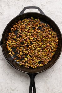 A black cast iron skillet filled with roasted pistachios sits on a light textured surface.